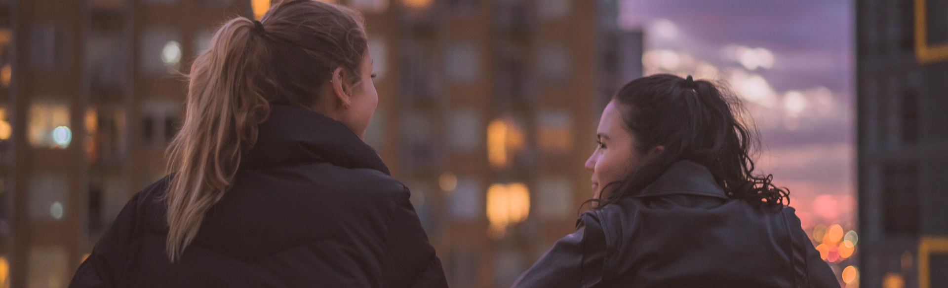 Two women talk to each other on a rooftop in a downtown setting